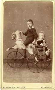 Young boy on rocking horse with young girl in wicker carriage behind, 1880s
